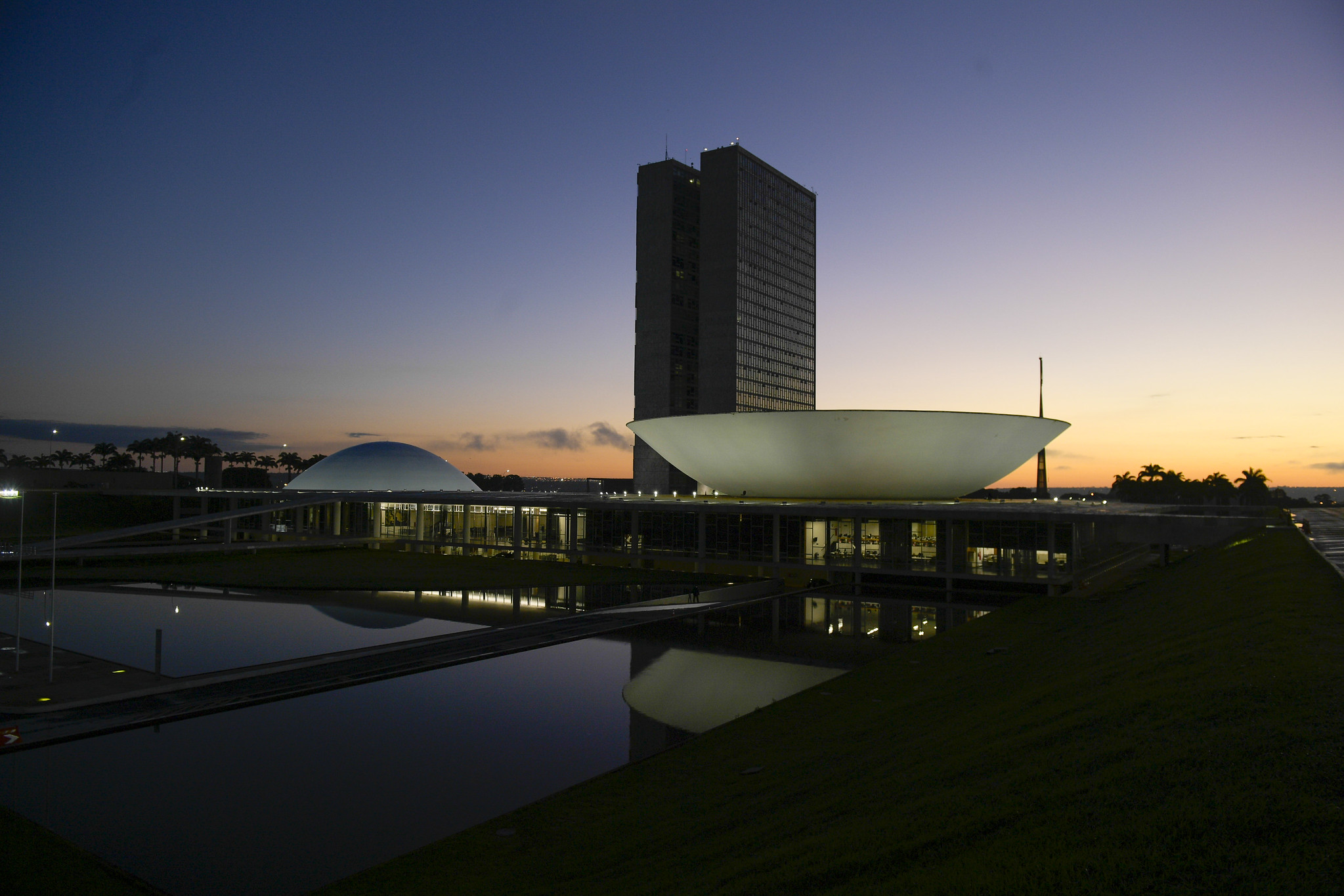 Edificações do Congresso Nacional em Brasília. A imagem dos prédios está refletida no espelho d'água enquanto o sol se põe.
